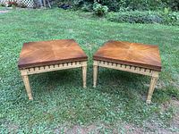 Photo of a pair of mid century side tables on grass showing square tops with a sunburst wood grain pattern and carved wooden legs with fluted detailing.