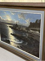 Framed landscape oil painting showing a nighttime coastal town illuminated under a cloudy sky, featuring waves, rocky shore, buildings, and a docked ship.