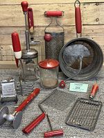 Photo showing assorted vintage red handled kitchen tools including graters, potato masher, whisks, flour sifter, and strainer with visible chippy paint and wear
