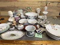 Overview photo of many assorted porcelain and ceramic items including plates, bowls, figurines, mugs and a lamp base on a table
