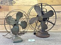 Two old fans shown side by side, one GE branded and one Kenmore, both metal with wire protective cages and heavily worn.