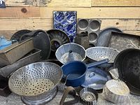 Large lot of assorted vintage baking pans, blue enamelware colanders, baking molds and metal utensils grouped against a wooden background.