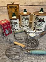 Vintage ceramic canisters labeled Tea, Coffee, Sugar with wooden lids and green handles with associated cooking utensils and vintage kitchen items in foreground.