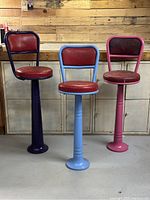 Three vintage metal swivel lunch counter stools with purple, blue, and pink frames and red vinyl seats and backs, arranged in front of wooden panel wall.