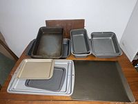 Photo shows assorted metal baking pans including rectangular and square cookie sheets and trays in silver and dark finishes.