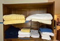 Folded assorted bathroom linen towels in yellow, white, blue, grey colors, stacked on a wooden shelf with metal hand towel stand in the middle.