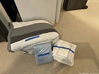 Folded and wrapped queen comforter with gray/white stripes next to packaged blue queen sheet set and packaged white king sheet set on carpet floor.