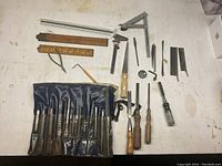 Photo showing all items arranged on a surface: 12 chisels in a roll-up case, various files and rasps, wooden and metal rulers, caliper, square, small hand plane, compass/divider, screwdriver, and drill bits.