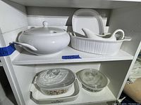 Clear glass Corning bakeware dishes with clear lids; one with floral pattern and one with vintage gold and black design, and white ceramic covered rectangular baking dish