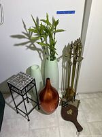 Photo showing fireplace tools set beside three vases (copper and green) plus a black metal plant stand with tile top and a decorative fireplace bellows.