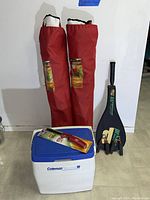 Two red folding chairs in carrying bags standing next to a blue and white Coleman cooler with a red utility knife on top and a black bag containing a badminton racket.