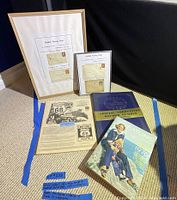 Photo showing two framed displays with four British penny postage stamps, Route 66 trip guide, Eatons catalogue 1976 and Illustrated London News 1953 coronation edition lying on carpeted floor