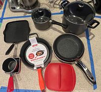Overview of assortment showing black pot with lid, small saucepan with red handle, grill pan, Lagostina frying pan with orange handle, and red silicone pan cover