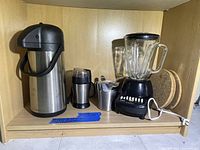 Photo showing a stainless steel coffee thermos, a coffee bean grinder, metal cream dispenser, black blender with glass jar, and two round cork trivets on a wooden shelf.
