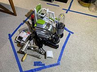 View showing multiple kitchen gadgets including hand mixer, spiralizer, egg beater, blender parts, and other utensils all arranged on carpeted floor with blue tape markings.