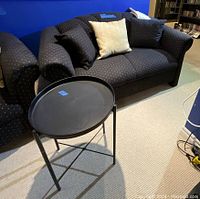 Photo showing the loveseat with dark upholstery featuring a white dot pattern, two cushions placed on it, and the black round metal side table beside it.