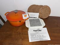Orange 12-inch cast-iron Dutch oven with lid, cork trivets, and stone heat retention plate on wooden table.