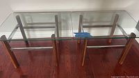 Pair of mid-century modern side tables with glass tops, teak legs, and chrome horizontal braces.