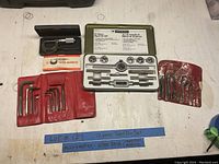 Photo of the 12-piece tap and die set in green metal case, micrometer in black case, and two rolled red fabric Allen key sets.