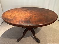 Full front view of the antique burled walnut oval table showing the wood grain and central pedestal base with four legs and castors.