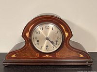 Front view of the wooden mantle clock showing the curved top, decorative inlay, and clock face inside glass cover.
