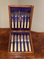 Front view of the wooden box open displaying 6 silver plated forks with ivory colored handles and six knives with matching handles inside purple velvet lining.