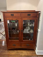 Front-facing view of the Mission Oak display cabinet with two glass-paneled doors, visible shelving, four drawers with metal handles, and sturdy oak construction.