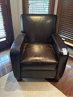 Full front view of the brown bonded leather armchair showing its padded armrests, cushioned seat and backrest, resting on wooden legs, situated near windows with blinds.