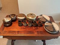 View of the entire pottery set on a wooden table showing arrangement of plates, bowls, cups, and a lidded pot in brown and cream glaze.