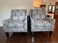 Pair of modern lounge armchairs side-by-side, showing abstract circular pattern upholstery in soft purples, green, and cream on a chenille-like fabric, dark wooden legs on hardwood floor