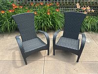 Pair of black resin wicker outdoor chairs set on a concrete patio with greenery and flowers in the background.