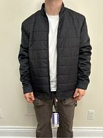 Front view of mens black quilted nylon jacket with black suede sleeves and white undershirt worn by model, showing full front with zipper closed and tags attached.