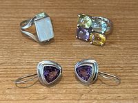 All three pieces displayed together on wooden surface: rectangular silver ring, multi-gemstone silver ring, and amethyst earrings.