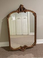Front view of vintage rectangular wall mirror with ornate gold gilded frame and carved details at top center and bottom. Reflects white closet doors and baseboard.