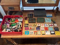 Wide overview photo showing a large red box filled with hundreds of matchbooks and various collectible tins on a wooden table, some matchbooks arranged neatly in rows to display vintage designs.