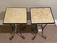 Pair of matching vintage wrought iron end tables with different tile tops, one with sun design and the other with decorative scroll pattern.