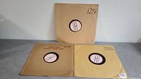 Three 16-inch records in plain cardboard sleeves with center label windows, arranged on gray surface.