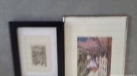 Two small framed artworks leaning against each other on a flat surface. One frame is silver with a colored city street scene, the other is black with a monochrome print.