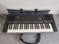 Front view of the Technics SX-K200 keyboard piano with all keys visible, placed on grey surface with carrying case behind.