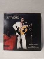 Close up of 'Paul Simon In Concert Live Rhythmin'' record album cover showing Paul Simon playing guitar on stage in white suit.