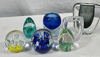 Six colorful blown glass paperweights featuring bubbles, swirls, and glitter inside.