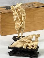 Two carved faux bone Far Eastern fisherman figures on black wooden bases with a wooden box in the background.