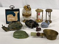 Overall view showing all items: two hourglass timers, pressed glass lighter, wood whale wall art, cast metal snail, carved frogs, clay whale, wax bunny, pottery beetle, brass silk iron.