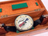 Compass device mounted in black metal gimbal structure inside wooden box with felt padding.