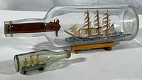 Photo showing both vintage ships in bottles side by side, larger one on wood stand and smaller one with fixed base inside bottle.