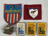Photo showing the embroidered Squaw Valley patch, Squaw Valley enamel brooch on red backing, three Lake Placid 1980 Olympic pins, and Innsbruck pin.