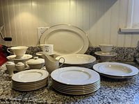 Overview of entire set with multiple plates stacked, serving pieces, and cups with saucers arranged on granite countertop.