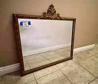 Full view of the antique ornate framed mirror leaning against a wall on a tiled floor, showing overall size and style.