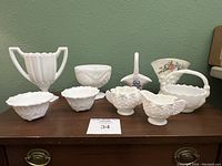 Nine assorted milk glass pieces on brown wooden surface against green wall, including bowls, baskets, and vases with floral and textured patterns.