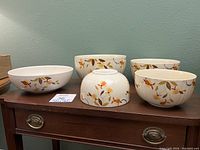 Five ceramic bowls with Autumn Leaf pattern, three ribbed and two smooth-sided, arranged on a wooden dresser.